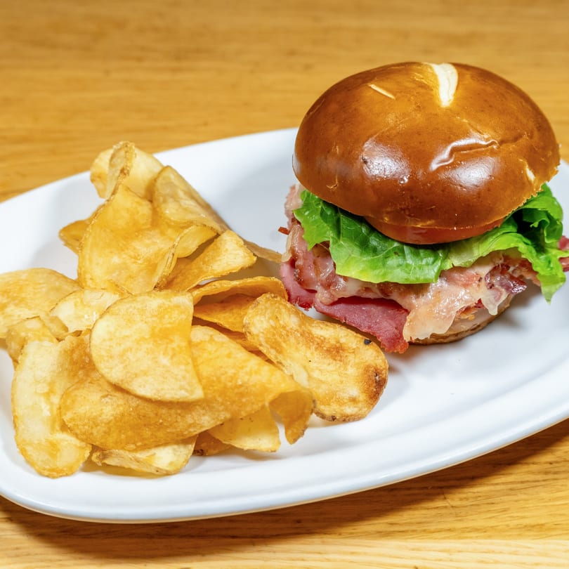 A pastrami sandwich on a pretzel bun with lettuce, tomato, and melted cheese, served with potato chips.