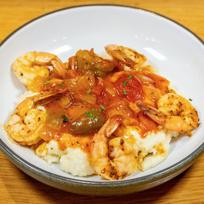 Shrimp and grits with a rich tomato and pepper sauce, garnished with chives in a white bowl.