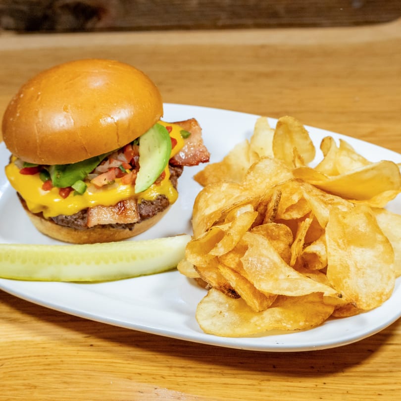 Cheeseburger with bacon, avocado, and pico de gallo, served with chips and a pickle spear.