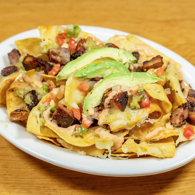 A plate of nachos with melted cheese, pork belly, guacamole, salsa, and avocado slices.