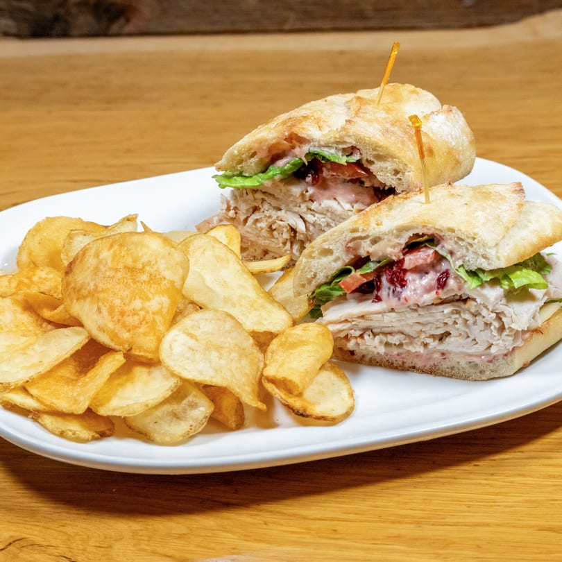 Turkey sandwich with lettuce, tomato, and cranberry sauce, served with potato chips on a white plate.